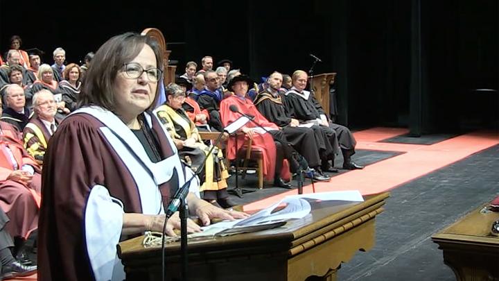 A woman stands at a podium, speaking into a microphone. She is wearing a maroon graduation gown.