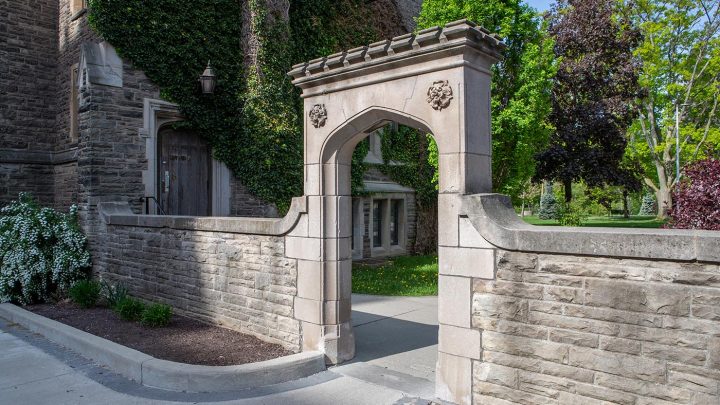 The sun shines on a stone archway in front of an old stone building on McMaster's campus.