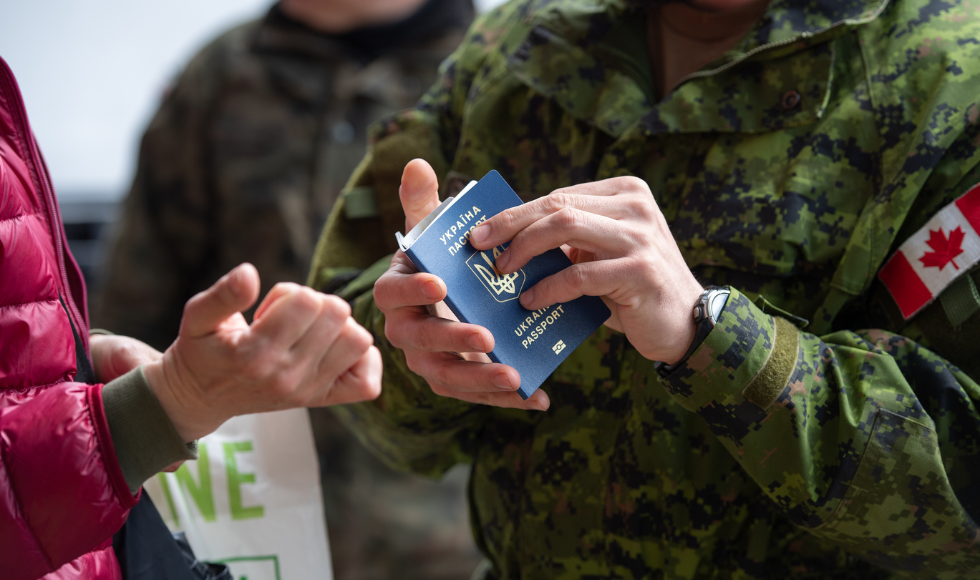 The hands of a person in Canadian military uniform holds an open passport as a person wearing a red jacket holds out their hands to take the passport.
