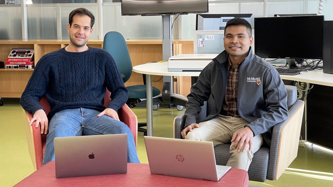 Two grad students sit on chairs with their laptops open. They are smiling.