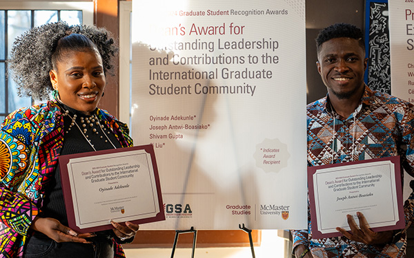 Two graduate students hold certificates as they stand on stage beside a Graduate Awards sign.